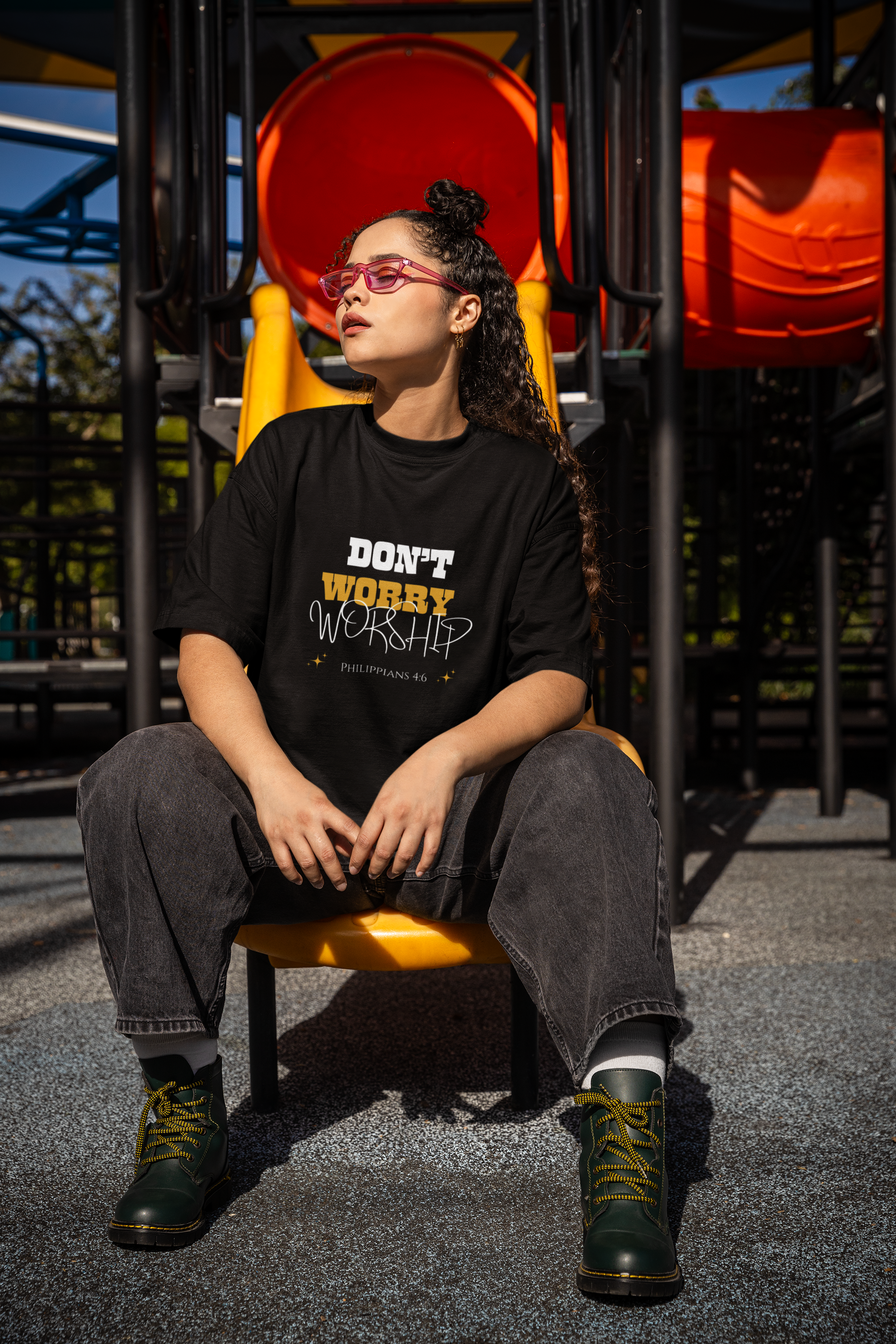 Girl with glasses sitting on a playground chair wearing a black t-shirt with dont worry worship text, wide legged jeans and boots